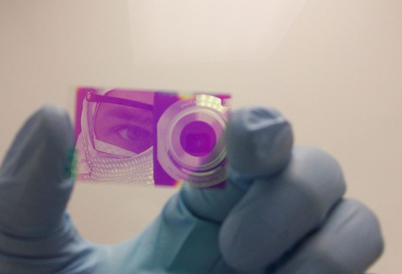 A gloved hand holds a glass slide coated in a magenta thin film. The
                    film reflects the person photographing it, in a mask and safety glasses.