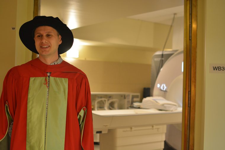 Mathieu Boudreau in McGill doctoral regalia, standing at the door of
                    the scanner suite at the McConnell Brain Imaging Centre, the 3 T MRI
                    visible through the window behind him.